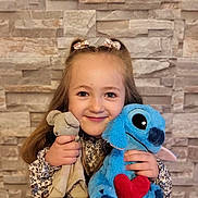 Giulia a rejoint le concours — aidez-le/la à gagner de superbes lots ! beige, blue, child, cute, girl, hairband, happy, holding, indoor, long_hair, patterned_clothing, person, plush_toy, portrait, red_heart, smiling, stone_wall, stuffed_animal, wall, young