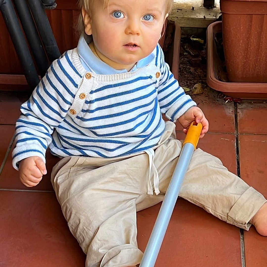 Tommaso participe au concours pour gagner de l'argent avec cette photo : baby, barefoot, beige_pants, blue_eyes, casual_clothing, child, curious, floor_tiles, indoors, long_object, person, plants, potted_plants, pottery, sitting, stick, striped_sweater, terracotta_floor, toddler, young_child