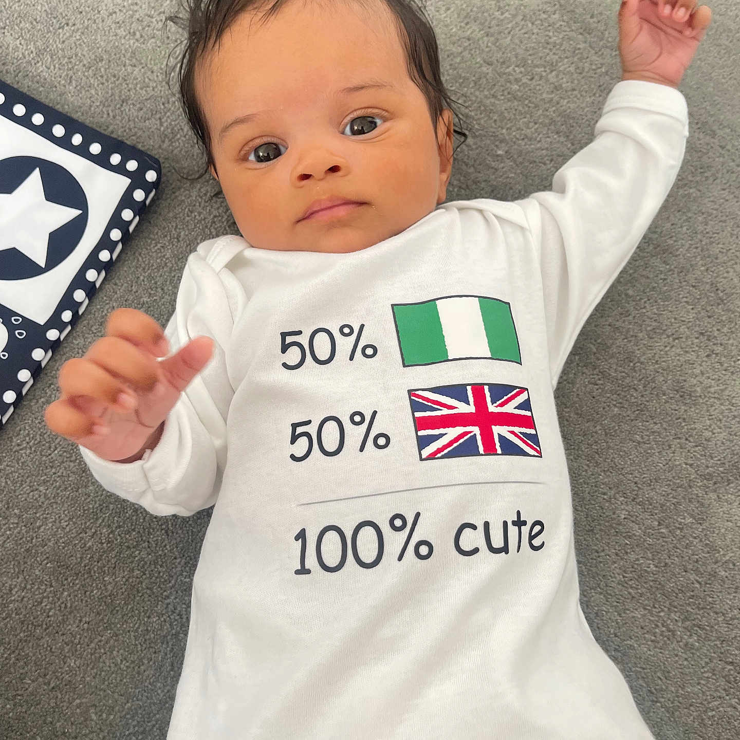 Exton joined the competition — help win amazing prizes! baby, carpet, child, clothing, cute, eyes, face, flag, hair, hand, indoors, infant, lying_down, nigeria_flag, onesie, soft_texture, text, toy, uk_flag, white_clothing