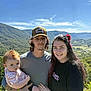 baby, man, woman, outdoor, mountain, sky, nature, family, portrait, smile, hat, greenery, child, casual_clothing, holding, sunlight, scenic, red_bow, long_hair, tattoo