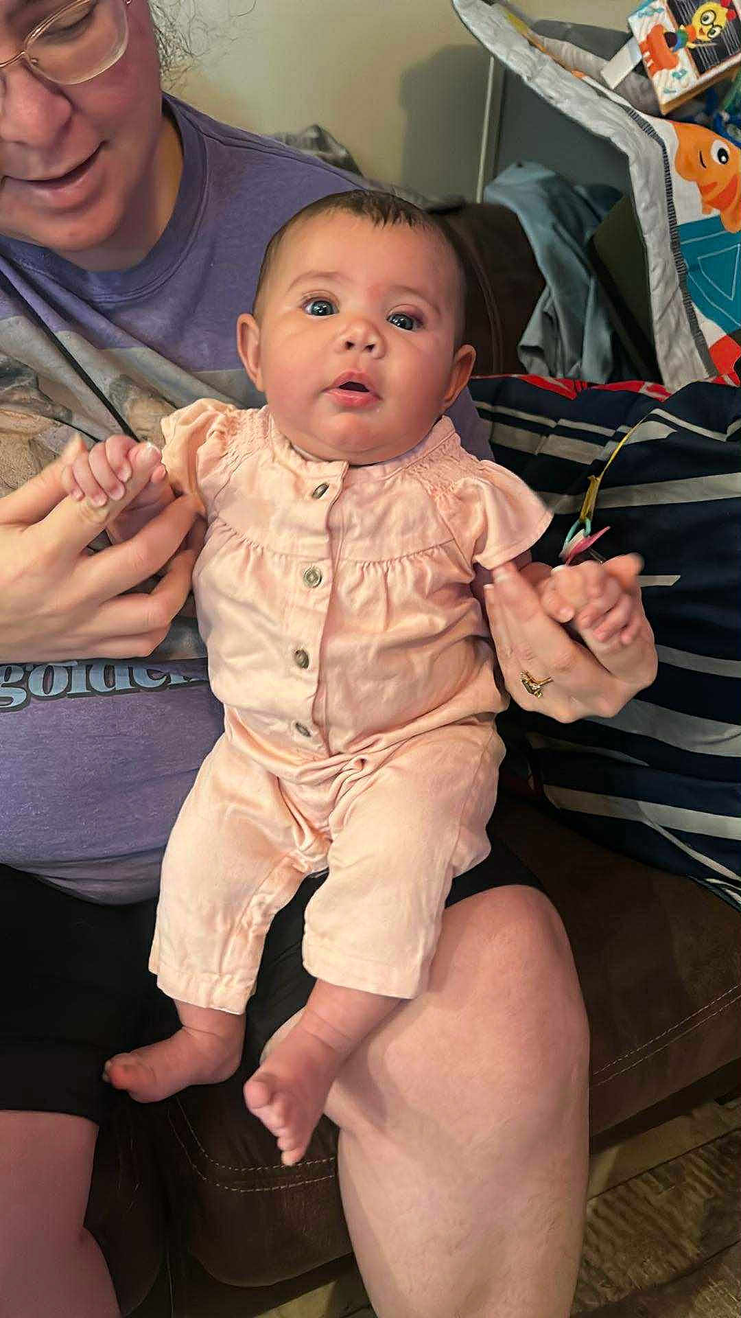 Armani is registered to the contest to win money with this photo: baby, infant, adult, glasses, purple_shirt, peach_outfit, couch, blanket, hand_holding, indoor, person, seated, cute, face, expression, skin, furniture, comfort, casual, home