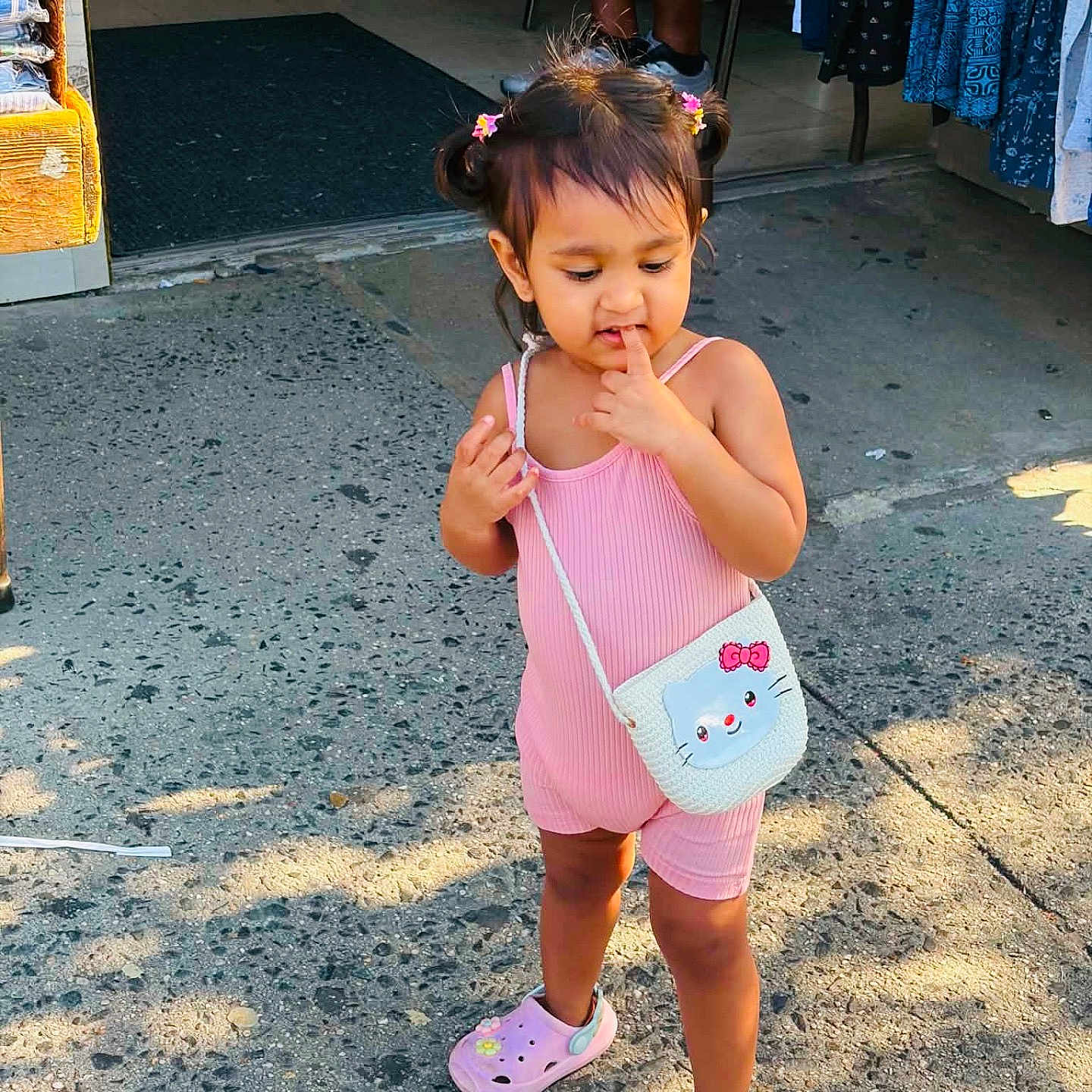 Anjuman is registered to the contest to win money with this photo: child, toddler, girl, outdoor, portrait, standing, smiling, pink_outfit, hair_bows, pigtails, handbag, cat_purse, pink_shoes, clogs, shop, storefront, sidewalk, pavement, sunlight, fashion