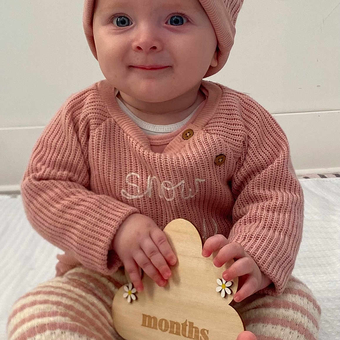 Inara joined the competition — help win amazing prizes! baby, barefoot, child, cozy, cute, feet, flower_shape, hands, happy, indoors, infant, knit_sweater, pink_hat, portrait, sitting, smiling, soft_lighting, striped_pants, wooden_sign, young_child