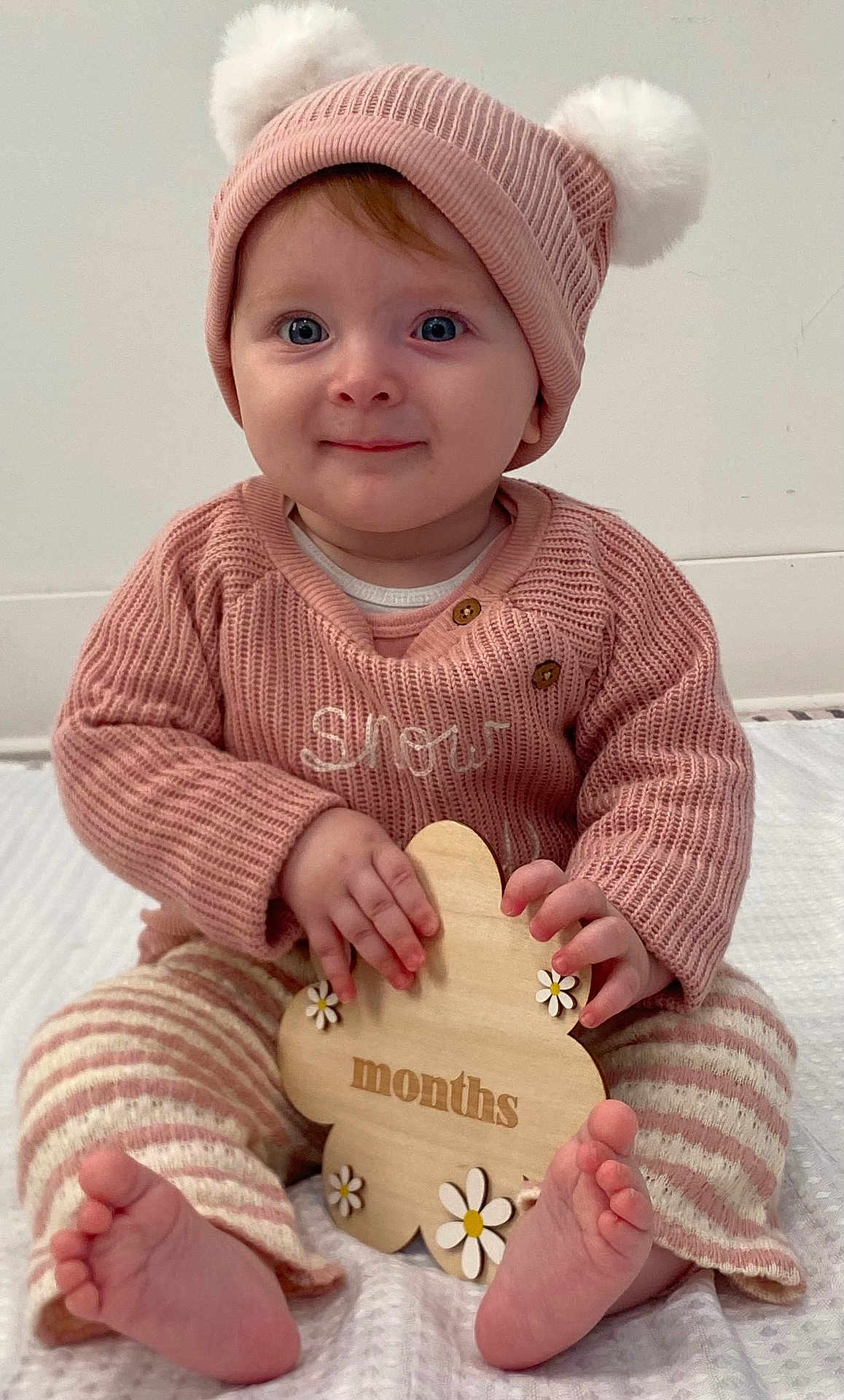 Inara joined the competition — help win amazing prizes! baby, child, infant, pink_hat, knit_sweater, striped_pants, barefoot, wooden_sign, flower_shape, smiling, sitting, indoors, cute, portrait, soft_lighting, cozy, happy, young_child, hands, feet