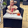 toddler, child, outdoor, wicker_chair, snack, sippy_cup, pigtails, cushion, patio, plants, fence, table, footwear, clothing, person, hair_bow, greenery, relaxing, daytime, cute