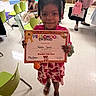 Winter is registered to the contest to win money with this photo: child, preschool_certificate, classroom, smiling, girl, dress, shoes, chairs, table, floor, indoor, education, celebration, achievement, happy, people, person, hair, skin, hand