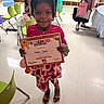 child, girl, certificate, preschool, classroom, smile, happy, dress, sandals, chairs, floor, education, celebration, achievement, indoor, people, hair, holding, person, background