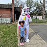 Winter is registered to the contest to win money with this photo: child, girl, costume, bunny, outdoor, sidewalk, grass, tree, building, fence, smile, happy, person, spring, playground, pink_shoes, floral_dress, yellow_bow, character, fun