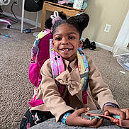 Winter is registered to the contest to win money with this photo: child, girl, smile, backpack, indoor, carpet, pigtails, clothing, snack, happy, person, furniture, table, room, casual, hair_accessory, toy, shoes, wall, power_outlet