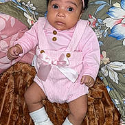 Nylah is registered to the contest to win money with this photo: baby, infant, pink_clothing, headband, bow, white_booties, knitwear, blanket, fur_blanket, floral_pattern, cushion, indoor, person, cute, wide_eyes, portrait, lying_down, child, soft_texture, cozy