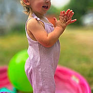 Riley is registered to the contest to win money with this photo: toddler, child, clapping, smiling, outdoor, splash_pool, water, balloons, playground, summer, pink_outfit, romper, barefoot, garden, backyard, portrait, side_profile, sunlight, colorful, happy
