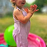 toddler, child, clapping, smiling, outdoor, splash_pool, water, balloons, playground, summer, pink_outfit, romper, barefoot, garden, backyard, portrait, side_profile, sunlight, colorful, happy