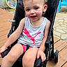 Riley is registered to the contest to win money with this photo: toddler, child, girl, smiling, sitting, stroller, outdoor, patio, deck, wooden_floor, hair_bows, blonde_hair, puma_shirt, sleeveless_shirt, sandals, playful, portrait, sunlight, pool_toy, brick_wall