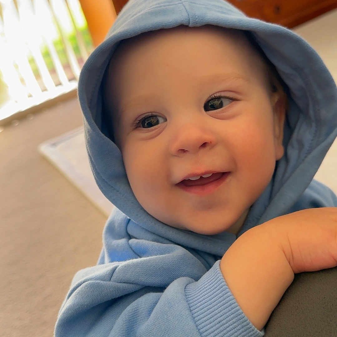 Luca joined the competition — help win amazing prizes! baby, blue_clothing, carpet, casual_clothing, child, cozy, curtains, cute, face, furniture, hand, happy, hoodie, indoor, portrait, relaxed, smile, soft_light, window, young_child