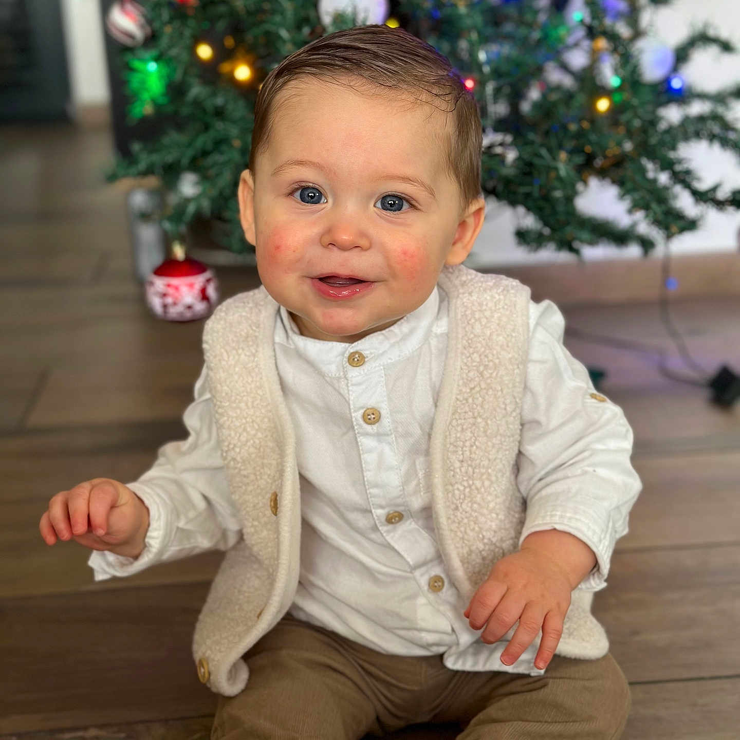 Ysao participe au concours pour gagner de l'argent avec cette photo : baby, beige_vest, blue_eyes, brown_pants, child, christmas_tree, cute, festive, happy, holiday, indoor, ornaments, portrait, rosy_cheeks, sitting, smile, toddler, white_shirt, white_shoes, wooden_floor
