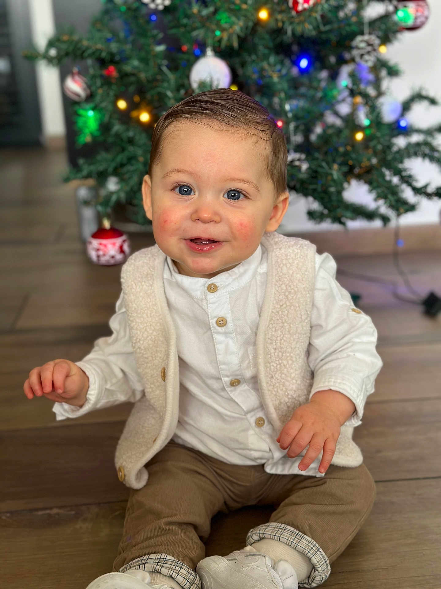 Ysao participe au concours pour gagner de l'argent avec cette photo : toddler, child, baby, smile, blue_eyes, rosy_cheeks, white_shirt, beige_vest, brown_pants, white_shoes, christmas_tree, ornaments, holiday, indoor, wooden_floor, festive, happy, sitting, cute, portrait