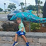 child, boy, smiling, sitting, stone_wall, dolphin_sculpture, outdoor, park, blue_shorts, sneakers, green_tshirt, plants, trees, car, sky, clouds, playful, daytime, public_space, decorative_art