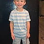 child, boy, smiling, striped_shirt, denim_shorts, beads, floor, refrigerator, indoor, wooden_floor, shorts, blue_shoes, happy, standing, casual_clothing, young, person, home, portrait, toy