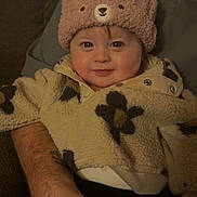 Lillie is registered to the contest to win money with this photo: baby, child, hat, bear_hat, fleece_jacket, floral_pattern, smile, cute, infant, person, portrait, warm_clothing, indoor, adult_arm, hand, cozy, seated, face, looking_at_camera, happy