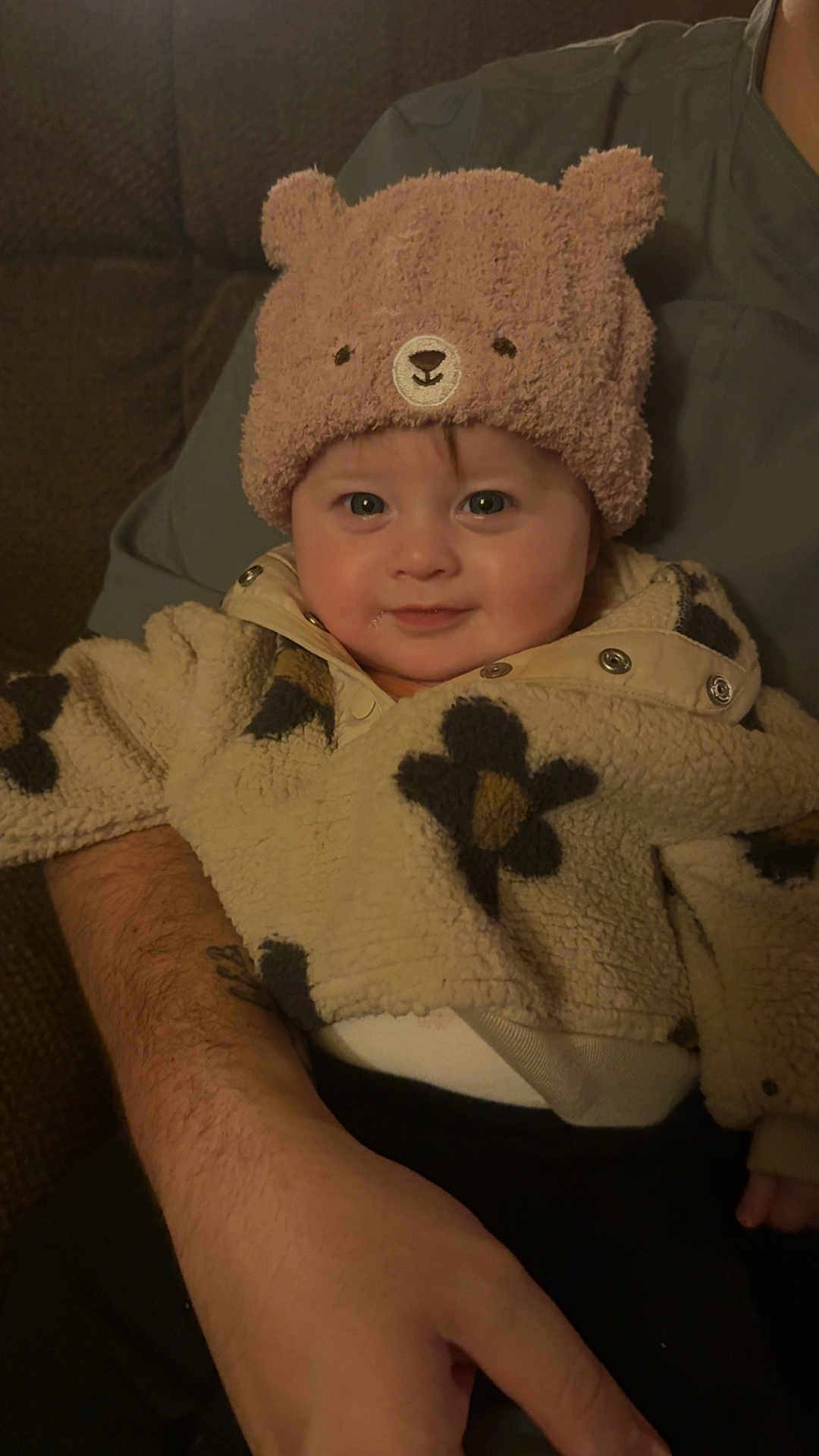 Lillie is registered to the contest to win money with this photo: baby, child, hat, bear_hat, fleece_jacket, floral_pattern, smile, cute, infant, person, portrait, warm_clothing, indoor, adult_arm, hand, cozy, seated, face, looking_at_camera, happy