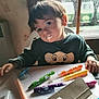 toddler, child, face, cream, markers, table, stickers, window, natural_light, curious, sitting, indoor, clothing, drawing, art_supplies, young_child, expression, hair, hand, cozy