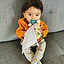 toddler, child, pacifier, plush_toy, orange_sweater, socks, gray_couch, indoor, cute, seated, curious, young_child, soft_toy, cozy, comfort, home, portrait, baby, furniture, casual_clothing