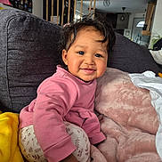 Kalyanne a rejoint le concours — aidez-le/la à gagner de superbes lots ! toddler, child, smiling, pink_sweater, patterned_pants, couch, blanket, cozy, indoor, home, furniture, happy, person, face, hair, living_room, plant, photography, cute, young_child