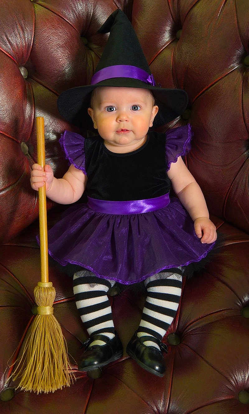 Amelia Rose joined the competition — help win amazing prizes! baby, child, costume, witch_hat, broomstick, striped_tights, black_shoes, purple_dress, leather_chair, portrait, cute, indoor, holiday, halloween, festive, sitting, young_child, face, smiling, playful