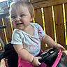 toddler, child, toy_car, pink, smiling, steering_wheel, hair_bow, outdoor, wooden_deck, white_shirt, skirt, happy, person, baby_teeth, playtime, summer, cute, hand, sunlight, portrait