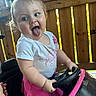 toddler, child, toy_car, pink_bow, blue_eyes, tongue_out, white_shirt, pink_skirt, steering_wheel, wooden_fence, outdoor, playful, cute, person, portrait, fun, happy, baby, daylight, smile