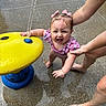 toddler, child, water_park, splash_pad, water_feature, yellow, pink_bows, swimsuit, wet, water_droplets, adult_hands, concrete, playing, summer, outdoor, fun, happy, person, barefoot, flip_flops