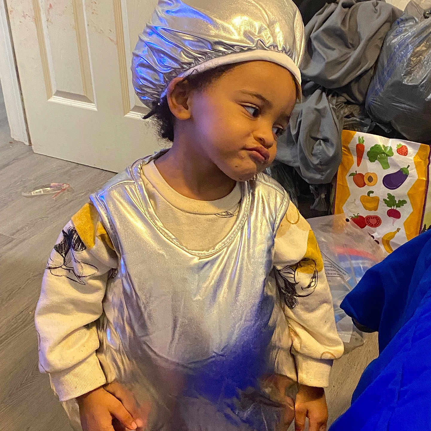 Clareice is registered to the contest to win money with this photo: brown_skin, casual, child, clothing, costume, expression, floor, grocery_bag, hat, household_items, indoor, long_sleeves, person, playful, short_hair, side_eye, silver, standing, toddler, wooden_floor
