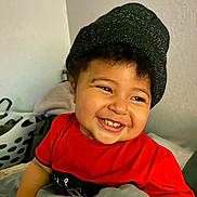 Clarence is registered to the contest to win money with this photo: beanie, blanket, casual_clothing, child, cozy, curly_hair, cute, expression, face, happy, hat, home, indoor, laundry_basket, person, portrait, red_shirt, smiling, teeth, toddler