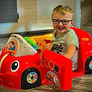 Evander is registered to the contest to win money with this photo: background, child, clothing, eyes, floor, furniture, glasses, happy, head, indoor, mouth, person, plastic_toy, playful, red, short_hair, sitting, smile, toddler, toy_car