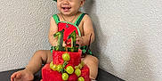 Matthew is registered to the contest to win money with this photo: baby, child, fruit_cake, watermelon, grapes, strawberries, smiling, birthday, hat, table, plate, celebration, cute, indoors, person, happy, food, cake, fruit, wall