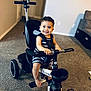 toddler, child, tricycle, indoor, carpet, smile, happy, person, toy, foot, handlebars, seat, wheel, clothing, black, white, room, furniture, living_room, bracelet