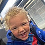 child, toddler, curly_hair, blue_jacket, smiling, teeth, indoor, bus_stop, window, seat, clothing, person, portrait, happy, casual, young, cute, spiderman_shirt, light, face