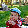 baby, child, blue_cap, red_tracksuit, grass, park, blanket, tongue_out, playful, sunny, trees, people, relaxing, outdoor, daytime, hand, smiling, nature, house, casual_clothing