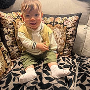 Adam participe au concours pour gagner de l'argent avec cette photo : baby, child, smiling, couch, cushion, floral_pattern, leaf_pattern, white_socks, yellow_vest, green_shorts, white_shirt, indoor, portrait, happy, seated, person, home, cozy, cute, infant