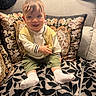 baby, child, smiling, couch, cushion, floral_pattern, leaf_pattern, white_socks, yellow_vest, green_shorts, white_shirt, indoor, portrait, happy, seated, person, home, cozy, cute, infant