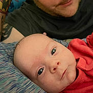 Serenity joined the competition — help win amazing prizes! baby, infant, person, adult, tattoo, face, skin, clothing, red_shirt, bed, blanket, closeup, portrait, eyes, expression, head, lying_down, indoor, parent, caring