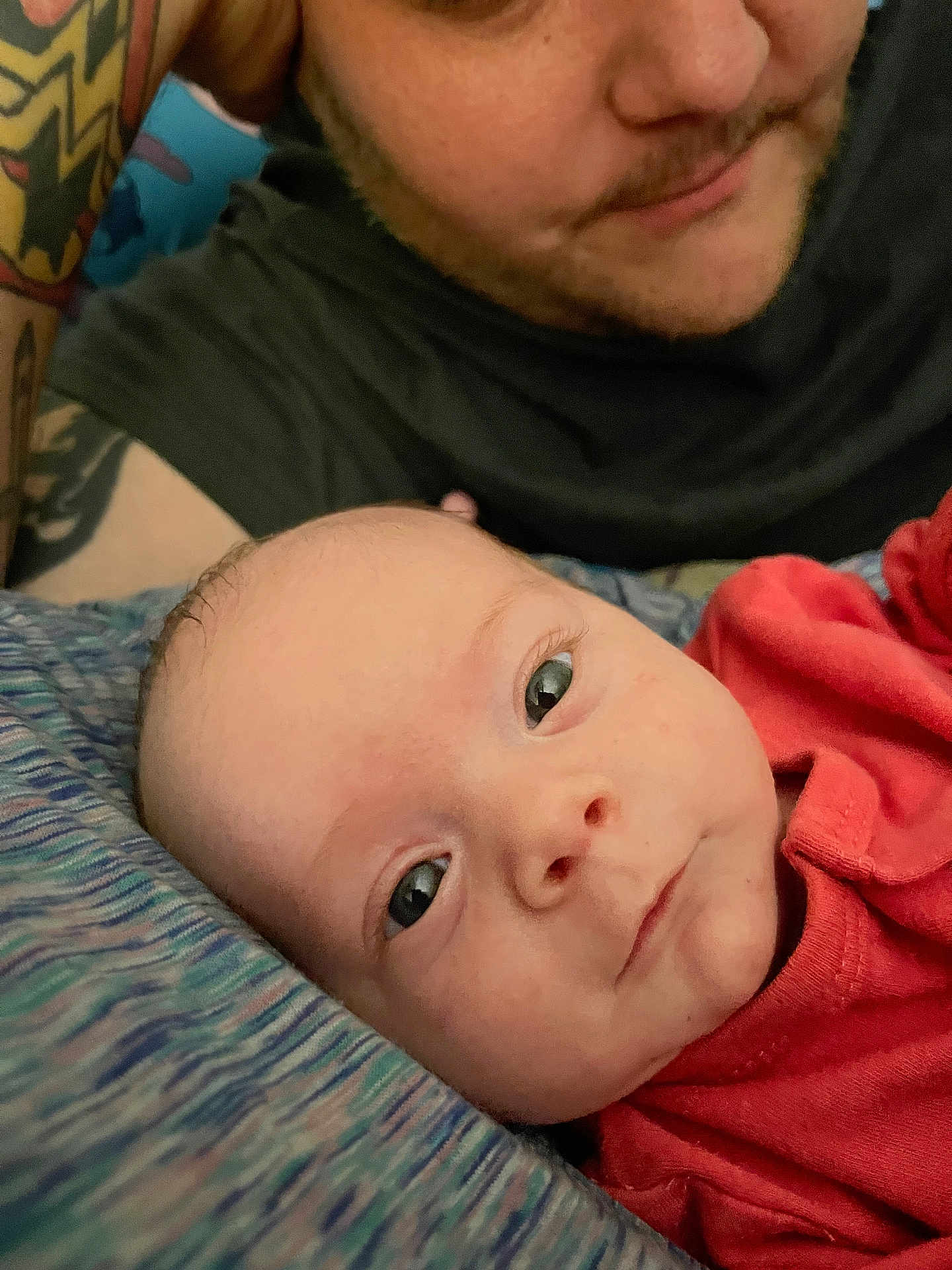 Serenity joined the competition — help win amazing prizes! baby, infant, person, adult, tattoo, face, skin, clothing, red_shirt, bed, blanket, closeup, portrait, eyes, expression, head, lying_down, indoor, parent, caring