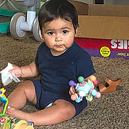 Jonah joined the competition — help win amazing prizes! toddler, child, toy, rattle, carpet, indoor, baby, clothing, footwear, cardboard_box, wipes, sitting, floor, person, playtime, plastic_toy, brown_sandal, shorts, shirt, curious