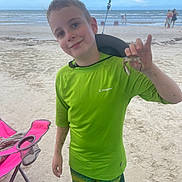 Logan is registered to the contest to win money with this photo: child, boy, beach, sand, ocean, sea, smile, green_shirt, hat, crab, chair, folding_chair, shorts, vacation, summer, shore, water, person, smiling, sunny