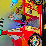 Oliver is registered to the contest to win money with this photo: toddler, child, arcade, racing_car, smiling, toy_car, indoor, colorful, playful, happy, shorts, tshirt, footwear, hand, seat, wheel, game, entertainment, light, reflection