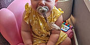 Scarlet is registered to the contest to win money with this photo: baby, pacifier, child, yellow_clothing, pink_chair, toy, plant, indoor, wood_floor, window, curious, sitting, cute, polka_dot, infant, small_hand, small_foot, face, blond_hair, home