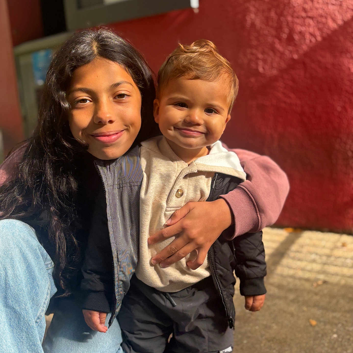 Aaron participe au concours pour gagner de l'argent avec cette photo : child, toddler, smiling, portrait, outdoor, sunlight, jacket, hoodie, jeans, sneakers, siblings, happy, cute, warm_clothing, person, standing, red_wall, candid, casual, daylight