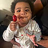 baby, child, lollipop, onesie, cherry_pattern, hand, face, smile, indoor, person, cute, skin, holding, infant, clothing, expression, adult_hand, young_child, portrait, closeup