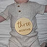 baby, infant, three_months, wooden_plaque, clothing, beige, quilt, lying_down, face, portrait, child, cute, indoor, plaid, casual, buttoned_shirt, young, skin, head, arms_outstretched