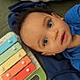 toddler, child, xylophone, toy, colorful, blue_clothing, face, eyes, curious, pillow, animal_print, indoors, laying_down, soft_surface, expression, young_child, playtime, music_toy, closeup, portrait
